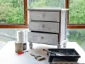 Mueble de tres cajones con acabado desgastado, listo para pintar muebles oscuros. Pinceles, rodillo y latas de pintura a su lado.