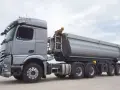 Srebrny, 3 osiowy samochód ciężarowy z naczepą typu wywrotka, gotowy do transportu dużej ładowności.