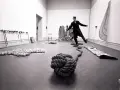 Un homme en costume, peut-être Lawrence Weiner, danse au milieu d'une installation artistique avec des cordes et des objets.