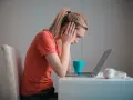 Zmartwiona kobieta przy laptopie, zastanawia się, czy porzucenie pracy wlicza się do emerytury. Na biurku kubek i słuchawki.