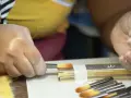 Une main tient un pinceau, prêt à peindre. D'autres pinceaux sont alignés à côté d'une règle.