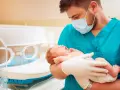 Lekarz w maseczce i rękawiczkach trzyma noworodka. To przykład troski w neonatologii, która zajmuje się opieką nad noworodkami.