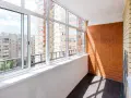 Wnętrze loggii z widokiem na miejską zabudowę. Ściany z cegły i tynku, przeszklony balkon z białymi ramami okiennymi.