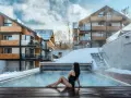 Kobieta w czarnym stroju kąpielowym siedzi przy podgrzewanym basenie. W tle nowoczesny hotel Karpacz w Karpaczu, otoczony śniegiem.