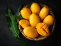 Koszyk pełen soczystych, żółtych mango, jedno przekrojone w kostkę. Obok zielone liście mango.