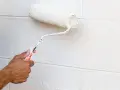 Main tenant, avis peinture carrelage sol : un rouleau blanc applique une nouvelle couche sur un mur en parpaings.