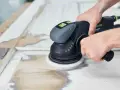 Mani che usano una levigatrice orbitale per rimuovere vecchia vernice e preparare il legno per una nuova finitura. Il processo per come lucidare il legno è in corso.