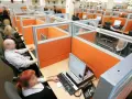 Wielostanowiskowe biuro, gdzie pracownicy obsługują telefony i komputery. W tle widać wiele przegródek, co może sugerować pra group nękanie.