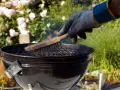 Mit einem Handschuh wird der Grillrost gereinigt. Rauch steigt auf, während die Bürste über die Roste fährt.