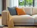 Sofá de tela beige con cojines azul y naranja, y una manta doblada. Ideal para limpiar sofá de tela y mantenerlo como nuevo.