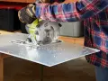 A person in a plaid shirt and gloves uses a circular saw to cut a sheet of plexiglass, creating small plastic shavings. This demonstrates that plexiglass is easy to cut with the right tools.