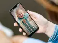 Ein lächelndes Baby im Strampler schaut aus dem Gitterbett. Eine Hand hält das Handy als Babyphone, um das Kind im Blick zu behalten.