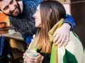 Pareja sonriente compartiendo una manta y una taza de té. Él le abraza por detrás. Ella le dice "muchas gracias en francés".