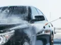 Man med t-grön tröja tvättar en svart bil med högtryckstvätt. Vatten och skum sprutar över bilen.
