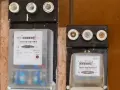 Dwa liczniki energii elektrycznej, jeden większy z czerwonym wskaźnikiem awarii, drugi mniejszy.