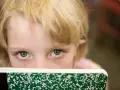 Oczy dziecka z blond włosami spoglądają zza zielonego zeszytu, ukrywając niepewność związaną z fobią szkolną.