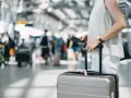 A traveler holds a passport and suitcase, mindful of carry-on size limits in a busy airport.