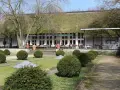 Das Teehaus im Englischen Garten Berlin mit seinem Reetdach und der Terrasse, auf der Gäste sitzen, ist von gepflegten Rasenflächen und Bäumen umgeben.
