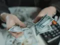 Dłonie liczą banknoty 100-dolarowe. Obok leży kalkulator, sugerując obliczanie kwoty, być może jako kara pieniężna za wyrządzoną krzywdę.