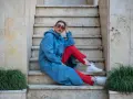 Kobieta w niebieskim płaszczu i czerwonych spodniach, w okularach, siedzi na schodach. Stylizacja to sportowa elegancja.