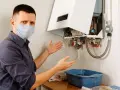 Uomo con mascherina mostra con le mani la caldaia a condensazione che perde acqua dal tubo di scarico, raccogliendola in una bacinella.