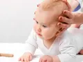 Dłoń delikatnie dotyka główki niemowlęcia, sprawdzając ciemiączko tylne. Obok leży drewniana zabawka.