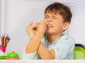 Junge mit zusammengepressten Augen und Händen vor dem Gesicht, ein Zeichen dafür, wie mit Tics bei Kindern umgehen. Buntstifte und eine Klingel liegen auf dem Tisch.