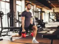 Mężczyzna wykonuje wykroki z kettlebellami, realizując swój plan treningowy na siłownię.