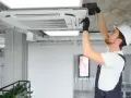 Technik w kasku montuje klimatyzator kasetonowy na suficie.