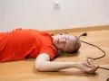 Mężczyzna w pomarańczowej koszulce leży na podłodze, obok kabel z wtyczką. Wygląda na to, że doświadczył porażenie prądem.