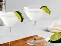 Dwa kieliszki z białym drinkiem, ozdobione plasterkami limonki. Obok miseczka z limonkami i shaker. Idealny kamikaze shot na gorący wieczór.