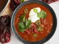 Un tazón humeante de chili con carne, adornado con crema agria, jalapeños y cilantro. Alrededor, chiles secos añaden un toque rústico.