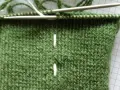 Grönt stickarbete med två vita markeringar, stickor och z tvinnat garn.