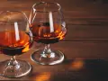 Dwa kieliszki z bursztynowym płynem, przypominającym bourbon lub whiskey, stoją na drewnianym stole.