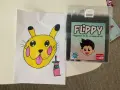 En gul Pikachu-liknande figur med rosa kinder och en tunga som sticker ut, bredvid en låda med färgpennor "FLIPPY" för dig som älskar att rita.