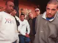 Un groupe de rap français des années 90/2000 pose devant un mur tagué. Ils portent des sweats et des jeans, typiques de l'époque.