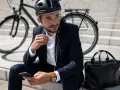 Mężczyzna w garniturze i kasku siedzi na schodach z rowerem w tle, słuchając muzyki przez słuchawki i przeglądając telefon.