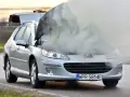 Srebrny samochód Peugeot z otwartą maską, z której wydobywa się gęsty dym.