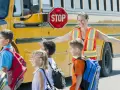 Des enfants montent dans un bus scolaire jaune, guidés par une adulte portant un gilet de sécurité orange.