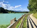 Sentiero sterrato con staccionata in legno lungo il lago di Tavon, circondato da alberi e montagne. Cielo azzurro con nuvole bianche.
