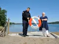 Två personer håller i en räddningsboj vid en sjö. En man i uniform och en kvinna ler. Trygg-Hansa vid Nörybyns camping.