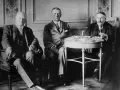 Tres hombres sentados, discutiendo los términos del **Tratado de Locarno**. Uno de ellos, a la derecha, manipula documentos sobre una mesa.