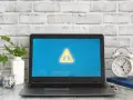 Laptop z symbolem ostrzeżenia na ekranie, obok budzik i rośliny.