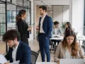 Il responsabile comunicazione discute con una collega in un ufficio moderno. Altri collaboratori lavorano ai loro tavoli.