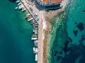 Widok z lotu ptaka na port z licznymi łodziami i jachtami cumującymi przy nabrzeżu. Woda jest krystalicznie czysta, a na dnie widać kamienie i ciemne plamy wodorostów.