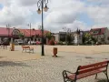 Rynek w Ślesinie z pomnikiem, ławkami i budynkami. Atrakcje tego miejsca zachęcają do odpoczynku.
