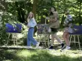 Trzy dziewczyny spacerują wśród obrazów na sztalugach. Jedna z nich, w niebieskich jeansach, przypomina młodszą wersję Tomasza Kłosowskiego.