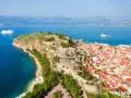 Widok na Nafplio w Grecji: miasto z czerwonymi dachami, twierdza na skale i biały statek wycieczkowy na lazurowym morzu.