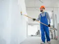 Maler mit blauer Latzhose und orangefarbenem Helm streicht eine Wand weiß. Informieren Sie sich über die malerarbeiten kosten für Ihr Projekt.