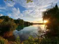 Sonnenuntergang am Rimsinger Baggersee. Die Wolken spiegeln sich im ruhigen Wasser, umrahmt von Bäumen und Schilf.